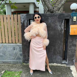 May include: A woman wearing a pink feather top and a matching pink skirt with a slit. She is wearing silver heels and sunglasses.