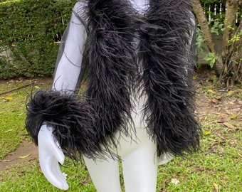 May include: A black feather jacket displayed on a white mannequin. The jacket features a sheer, light-colored fabric base, with dense black feathers trimming the collar, cuffs, and front. The mannequin is positioned outdoors on a grassy surface.