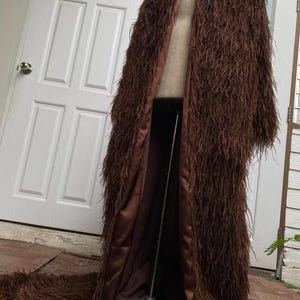 May include: A floor-length brown coat with a shaggy, textured exterior. The coat has a satin-like lining and a long train. The garment is displayed on a mannequin, showcasing its full length and design.