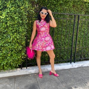 May include: A sleeveless, hot pink dress with floral embroidery. The dress is paired with matching pink heels and a feathered clutch. The woman is wearing white sunglasses.