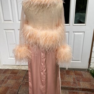 May include: A long, pink satin robe with a sheer, feather-trimmed overlay. The robe has long sleeves and a shawl collar.