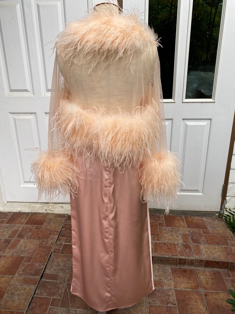 May include: A long, pink satin robe with a sheer, feather-trimmed overlay. The robe has long sleeves and a shawl collar.