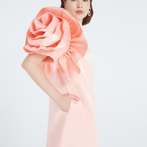 Könnte beinhalten: Ein rosa Kleid mit einem großen, gerafften Rosen-Detail auf einer Schulter. Das Kleid hat eine schlichte, A-förmige Silhouette und eine Tasche an der Seite.