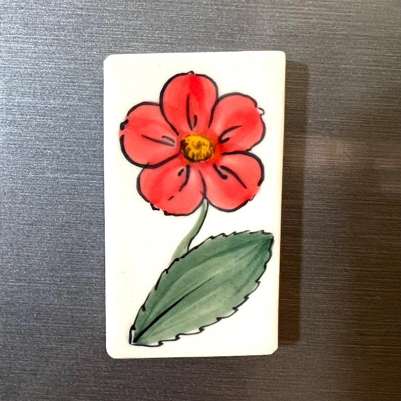 May include: A white ceramic tile with a hand-painted red flower with a yellow center and green leaves.