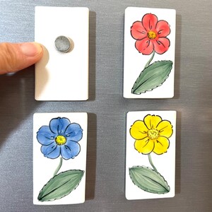 May include: Four rectangular ceramic magnets with painted flower designs. One magnet is blank. The flowers are red, yellow, and blue.