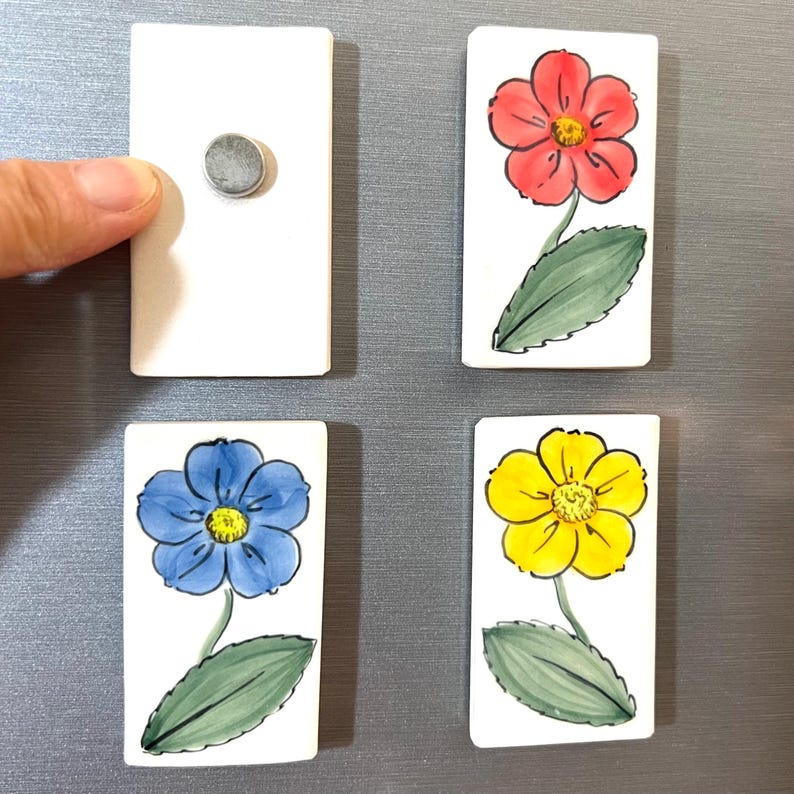 May include: Four rectangular ceramic magnets with painted flower designs. One magnet is blank. The flowers are red, yellow, and blue.