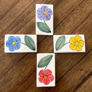 May include: Four ceramic tiles with hand-painted floral designs. Each tile features a different colored flower with green leaves. The flowers are purple, blue, yellow, and red.