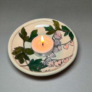 May include: A small, round ceramic candle holder with a lit tea light candle. The holder is decorated with green leaves and pink and purple floral designs. The candle holder is off-white with a thin black border.
