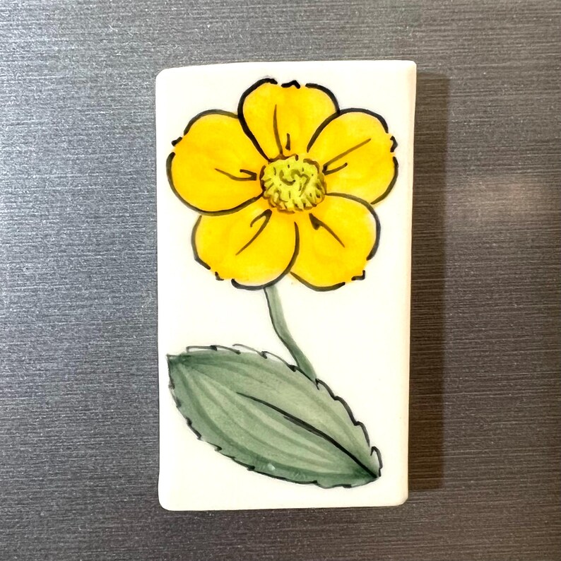 May include: A white ceramic tile with a hand-painted yellow flower with a green leaf. The flower has a yellow center and five petals.