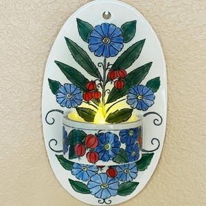 May include: A white ceramic wall sconce with a floral design. The oval-shaped sconce features blue flowers, green leaves, and red berries. A cylindrical candle holder is at the bottom, with a lit candle inside. The sconce has a hole at the top for hanging.