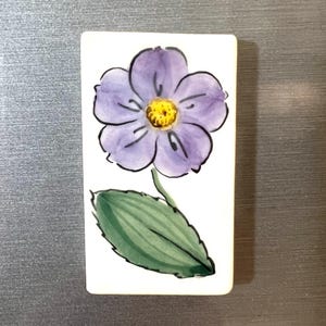 May include: A white ceramic tile with a hand-painted purple flower with a yellow center and green leaves.