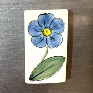 May include: A white ceramic tile with a hand-painted blue flower with a yellow center and a green leaf.