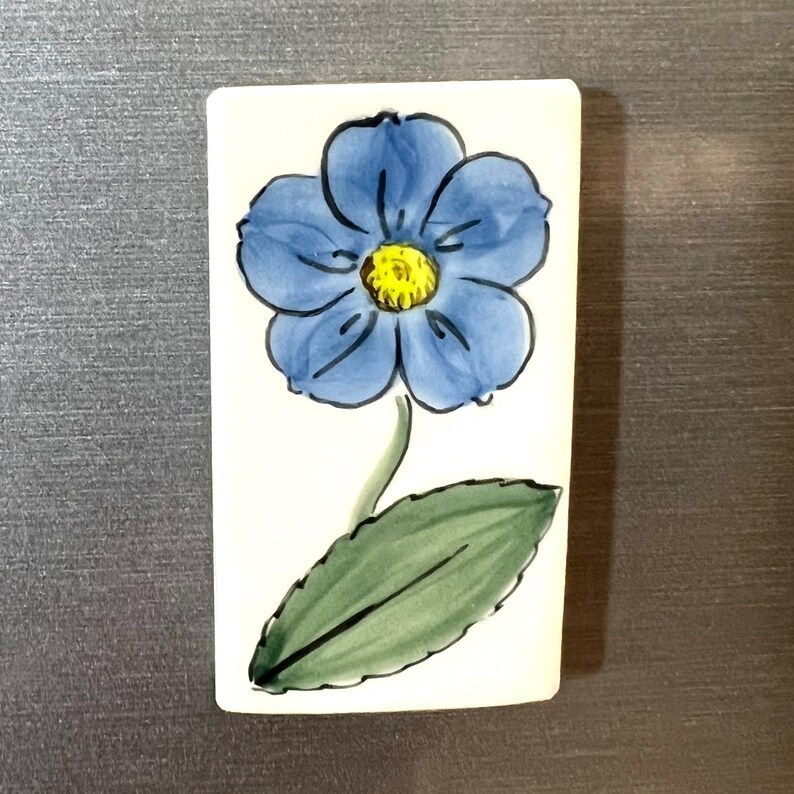 May include: A white ceramic tile with a hand-painted blue flower with a yellow center and a green leaf.