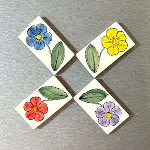 May include: Four ceramic tiles with painted flower designs. Each tile features a different colored flower: blue, yellow, red, and purple. The flowers are painted on a white background with green leaves.