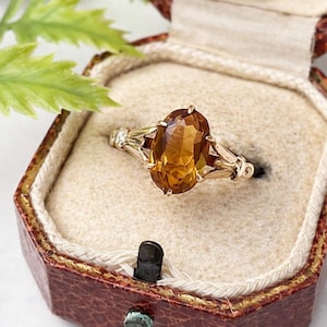 May include: A gold ring with a large, oval-cut, amber-colored gemstone. The ring is set in a vintage-style setting with smaller accent stones. The ring is displayed inside a vintage, red jewelry box with a cream-colored interior.