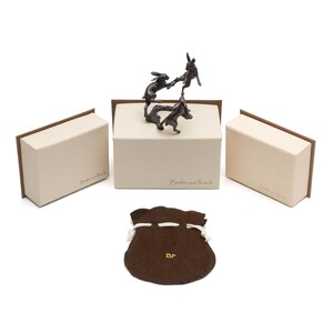 May include: A bronze sculpture of three hares in a playful pose, displayed on a white box with the text "Butler and Peach". The sculpture is accompanied by a brown velvet pouch with the initials "BP" embroidered on it.