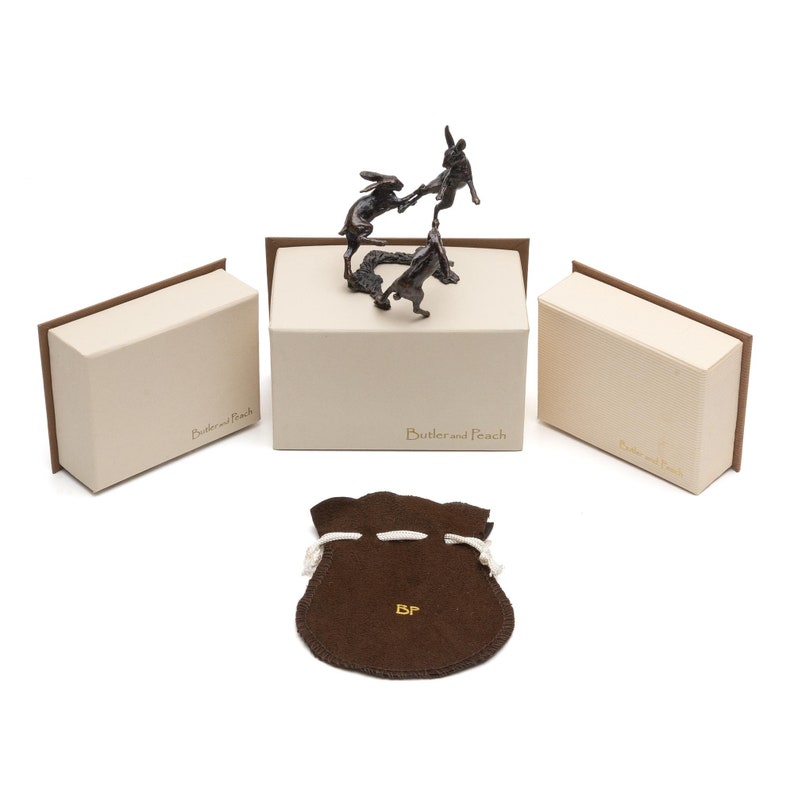 May include: A bronze sculpture of three hares in a playful pose, displayed on a white box with the text "Butler and Peach". The sculpture is accompanied by a brown velvet pouch with the initials "BP" embroidered on it.