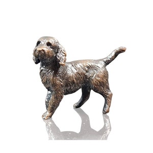 Puede incluir: Una figura de perro de color bronce con pelaje texturizado y orejas caídas. El perro está de pie con la cola ligeramente levantada. La escultura tiene una base reflectante, lo que sugiere una superficie pulida.