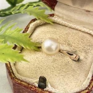 May include: A gold ring with a large, round pearl set in a vintage jewelry box. The box is red and cream-colored, with a cream-colored interior. Green leaves are in the background.