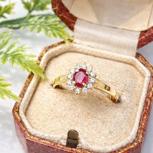 Può includere: Un anello d'oro con una pietra preziosa centrale di rubino ovale, circondata da un alone di piccoli diamanti rotondi. L'anello è incastonato in uno stile vintage e presentato in un piccolo portagioie antico.