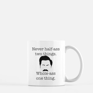May include: White ceramic mug with a black graphic of a mustache and hair, with the text "Never half-ass two things. Whole-ass one thing." The mug has a curved handle and is suitable for hot or cold beverages.
