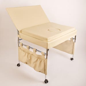 May include: Cream-colored massage table with a padded surface and a face cradle. The table has a chrome frame, storage pockets, and rolling wheels for easy movement. The backrest is adjustable.