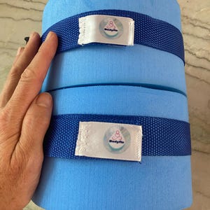 Op de afbeelding: Een lichtblauwe zwemhulp van schuim met twee donkerblauwe banden. Elke band heeft een wit label met de tekst "Bodyflo" en een logo. De hulp is cilindrisch en lijkt gemaakt van een zacht, drijvend materiaal.