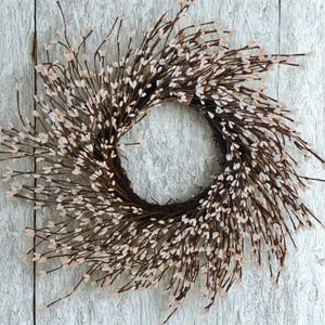 May include: A circular wreath made from brown twigs, decorated with small, pale pink, oval buds. The wreath is set against a white, weathered wooden backdrop, giving a rustic and natural feel. This decorative wreath is suitable for seasonal home decor.