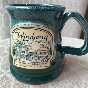 May include: A dark green ceramic mug with a speckled glaze finish. The front features a cream-colored oval label with a black line drawing of a building and the text: Windsong A Mountain Inn, Great Smoky Mountains, North Carolina.