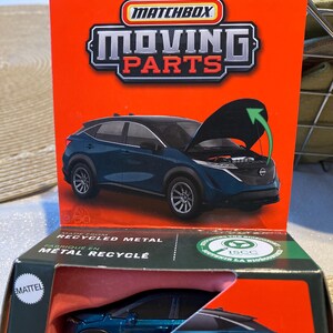 May include: A Matchbox Moving Parts die-cast model of a 2023 Nissan Ariya in a teal color. The car's hood is open, revealing the engine. The packaging is orange and black, with the Matchbox logo and the Nissan logo visible.