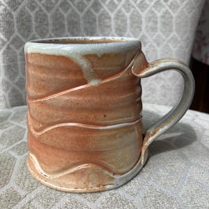 May include: Handmade ceramic mug with a unique, textured design. The mug features a warm, earthy color palette with shades of peach and cream. The handle is curved and sturdy, and the rim has a subtle, crackled glaze.