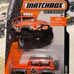May include: Matchbox Dune Dog toy car on a mission. The orange and gray vehicle has large black tires and a roof rack. The packaging includes the text "Guaranteed for Life" and a choking hazard warning.