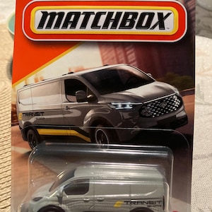 May include: Matchbox Ford Transit van toy in a gray and yellow color scheme. The packaging is orange and red with the Ford logo and the Matchbox logo. The text "NEW NOUVEAU 2025" is visible on the packaging.