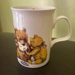 May include: White ceramic mug featuring a charming illustration of two teddy bears. One bear is brown, the other is yellow, and both are depicted in a friendly embrace. The mug has a curved handle and a slightly flared rim.