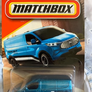 May include: A Matchbox toy car package featuring a blue Ford E-Transit van. The package has the Ford logo and the Matchbox logo. The van has black wheels and the text "E TRANSIT" on the side. The package is red and yellow.