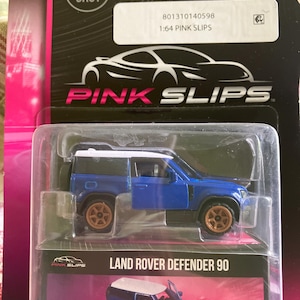 May include: A blue and white Land Rover Defender 90 die-cast model car in a clear plastic package. The model has gold-colored wheels and the door is open. The packaging includes the text "PINK SLIPS".