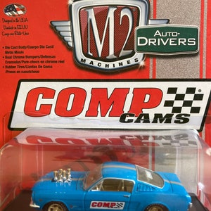 May include: A blue 1966 Ford Mustang Fastback diecast model car with a black and white COMP CAMS logo on the side. The car has a black interior and a chrome bumper. The car is on a red and white display base with the words "COMP CAMS" in black and white.