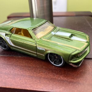 May include: A green die-cast toy car with black wheels and a white stripe along the side. The car has a yellow interior and a silver accent on the hood. The toy car is a miniature replica of a classic muscle car.
