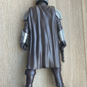 May include: A brown and silver action figure of a character wearing a helmet and a cape. The figure is holding a blaster in its right hand.