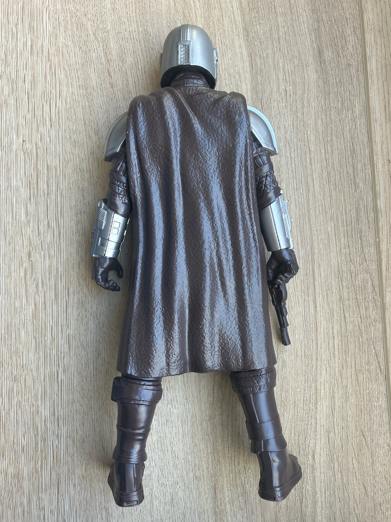 May include: A brown and silver action figure of a character wearing a helmet and a cape. The figure is holding a blaster in its right hand.