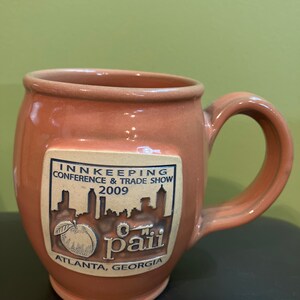 May include: A terracotta-colored ceramic mug with a beige-colored logo. The logo reads "Innkeeping Conference & Trade Show 2009" and "O.M. Paiii Atlanta, Georgia". The logo also features a peach and a cityscape.