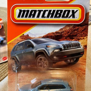 May include: A Matchbox Jeep Cherokee toy car in a light blue color, packaged on a red and orange card. The card features the Jeep logo and the Matchbox logo. The background shows a desert landscape.