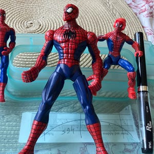May include: Three Spider-Man action figures in red and blue costumes, with a black pen. The figures are in various poses, with the largest one standing in the center. The pen is black with silver accents and has the text "0.7 mm" and "R2 inc."