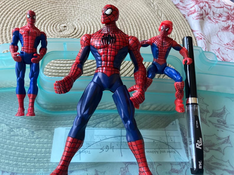 May include: Three Spider-Man action figures in red and blue costumes, with a black pen. The figures are in various poses, with the largest one standing in the center. The pen is black with silver accents and has the text "0.7 mm" and "R2 inc."