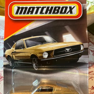 May include: A gold Ford Mustang die-cast toy car in its original Matchbox blister pack packaging. The card features the Matchbox logo, the Ford logo, and the number 61/125. The car has black wheels and a metallic gold finish, displayed in a clear plastic bubble.