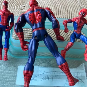 May include: Three Spider-Man action figures in dynamic poses. The figures are primarily red and blue, with detailed webbing patterns. One figure is viewed from the back, showcasing the spider emblem. The figures are on a light blue surface.