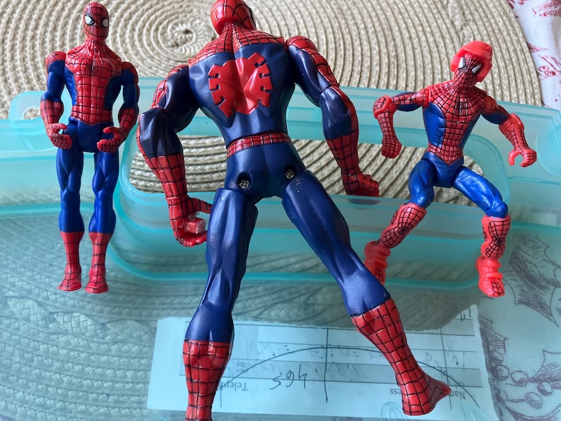 May include: Three Spider-Man action figures in dynamic poses. The figures are primarily red and blue, with detailed webbing patterns. One figure is viewed from the back, showcasing the spider emblem. The figures are on a light blue surface.