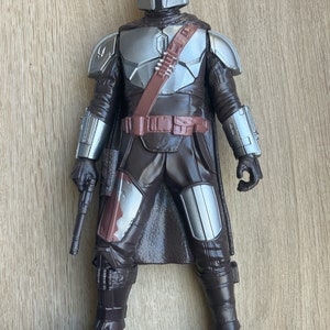 May include: A brown and silver action figure of a Mandalorian warrior. The figure is wearing a helmet with a T-shaped visor, a brown cape, and a silver chest plate. The figure is holding a blaster rifle in its right hand.