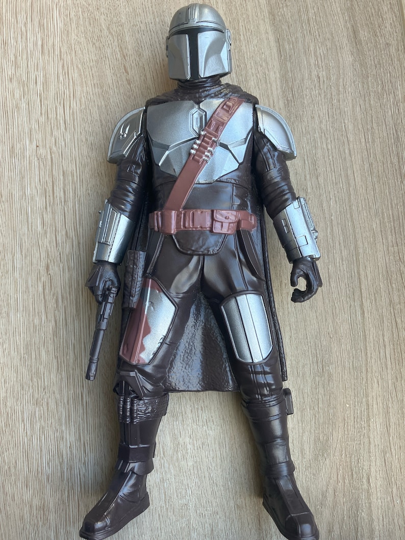 May include: A brown and silver action figure of a Mandalorian warrior. The figure is wearing a helmet with a T-shaped visor, a brown cape, and a silver chest plate. The figure is holding a blaster rifle in its right hand.