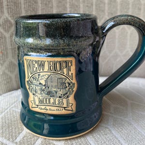 May include: A green ceramic mug with a brown and white label that reads "New Hope Mills Quality Since 1823". The label features a black and white illustration of a watermill.